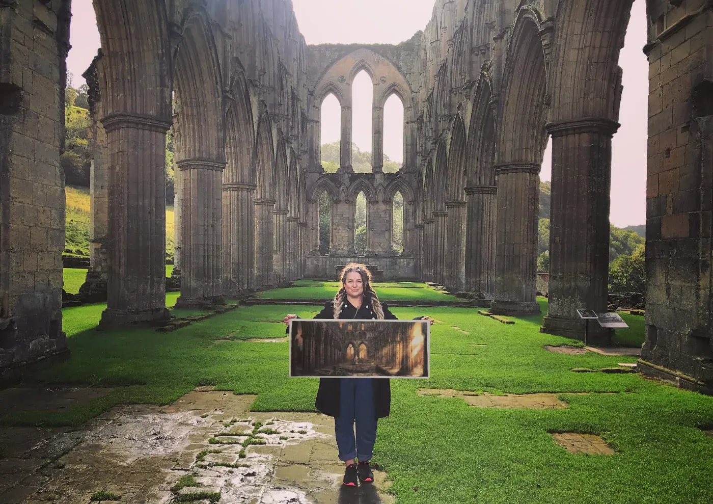 Juliana Loomer standing in a stone abbey ruin holding a large framed painting